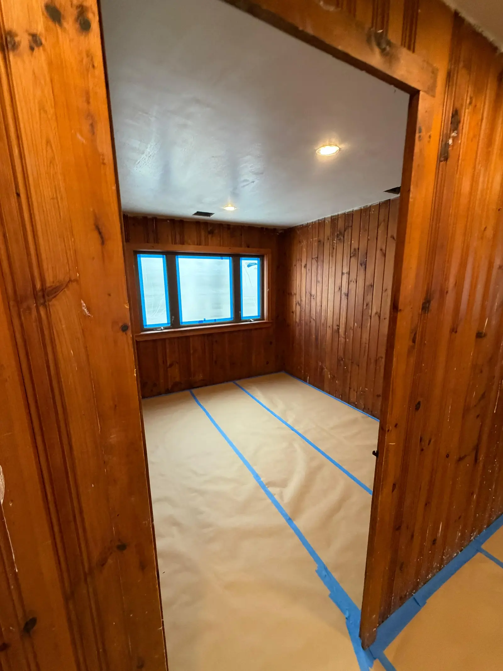 Bedroom with knotty pine paneling before painting, floor covered with paper and blue tape