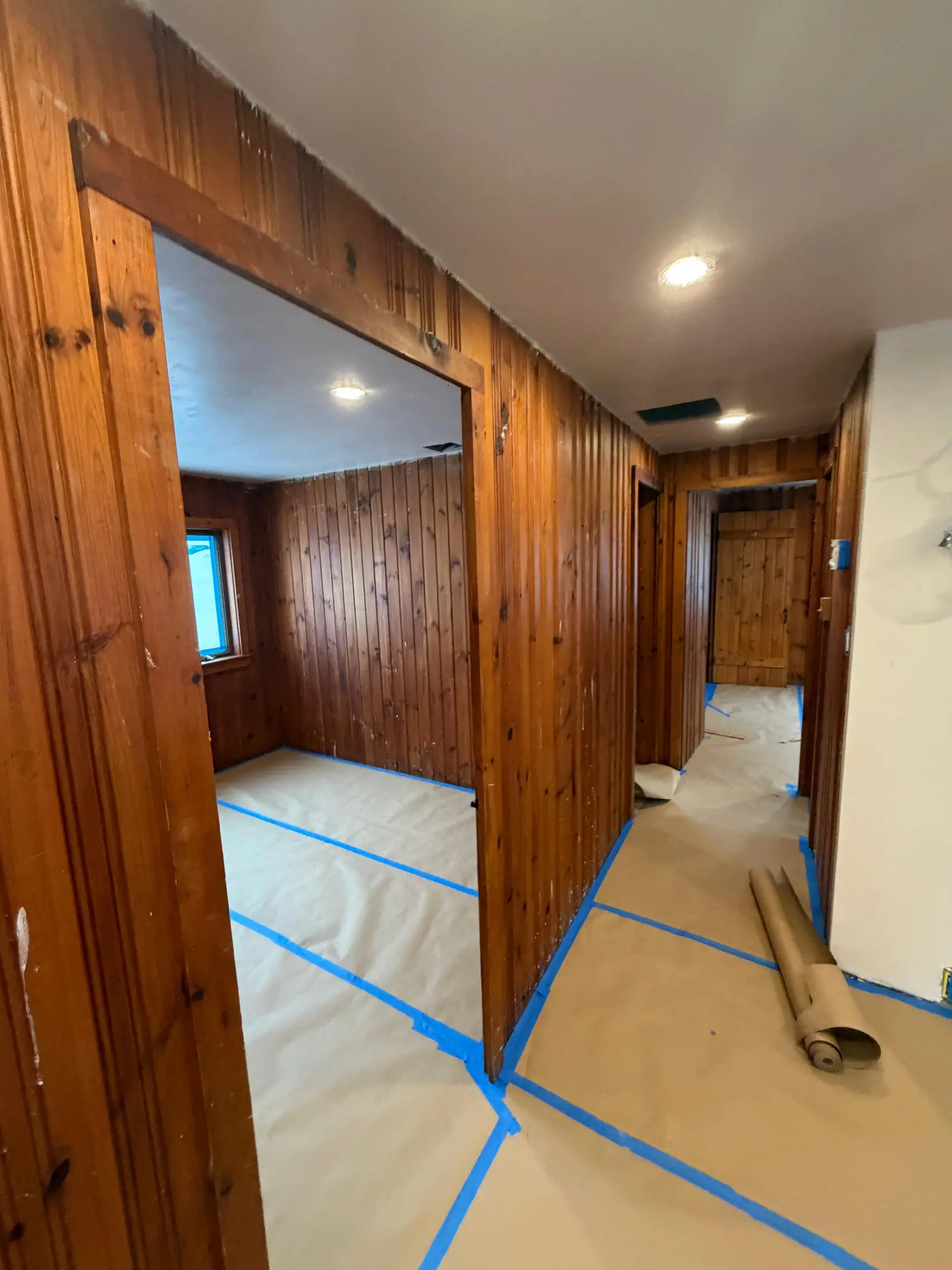 Hallway and adjoining room with knotty pine paneling, floors masked before painting