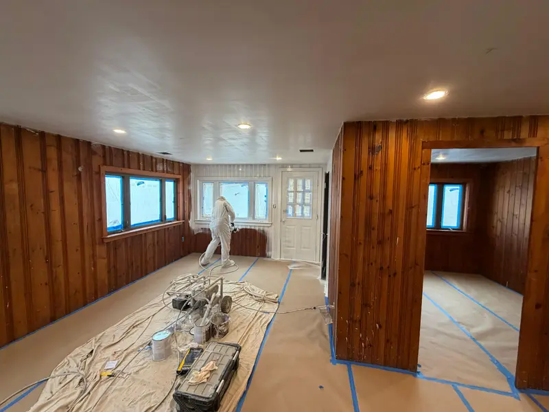 Painter in protective suit spraying primer on knotty pine paneling with floors and windows masked