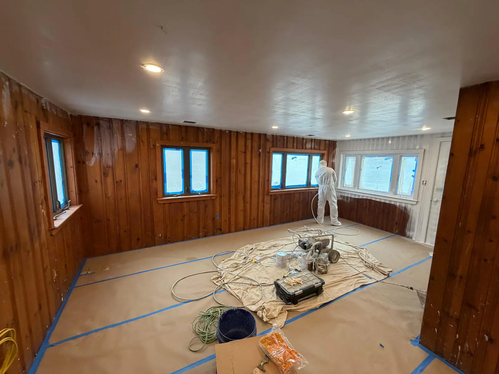 Wide view of a pine-paneled room being sprayed, with airless sprayer and paint cans on drop cloths